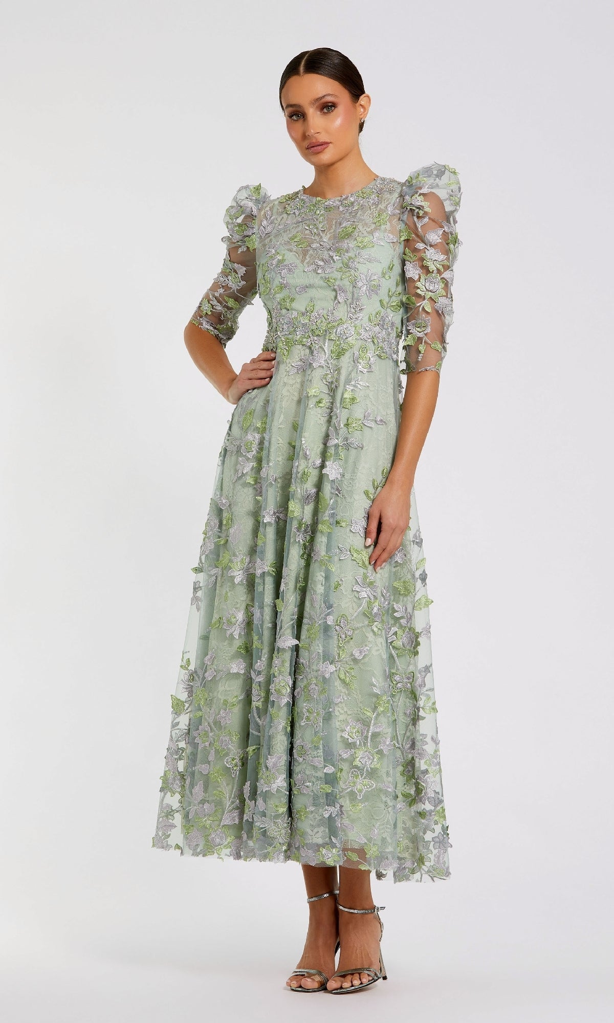 A woman stands against a plain white background wearing the Mac Duggal 68193 Tea-Length Lace Formal Dress in light green with floral embroidery, puffed sleeves, and strappy heels.